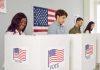 People voting in booths with American flag backdrop.