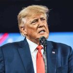 Trump Gifts Argentina $20BN – For What? Man in suit speaking at a microphone.