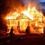 House engulfed in flames with firefighters present.