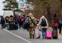 REFUGEE Aid SLASHED — Americans Next! Group of people carrying luggage walking along a road with a soldier