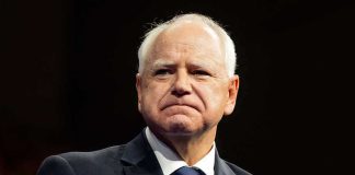 Walz DEFLECTS Massive Fraud With Race Card Man in suit with a U.S. flag pin.