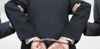 Person handcuffed in suit, escorted by two others.