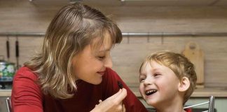 A mother playfully feeding her son in a kitchen filled with fruits and snacks