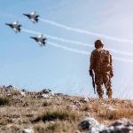 Soldier standing on rocky hillside watching fighter jets fly overhead