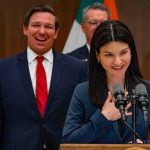 Ron DeSantis smiling behind a woman speaking at a podium