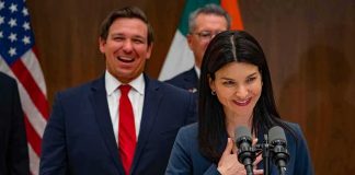 Ron DeSantis smiling behind a woman speaking at a podium