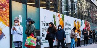 People wearing masks line up outside a store on a city sidewalk