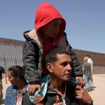 Man carrying a child in a red hoodie on his shoulders near a border fence