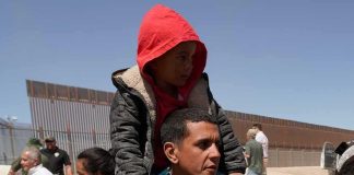 Man carrying a child in a red hoodie on his shoulders near a border fence