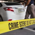Yellow crime scene tape in front of a white car with investigators nearby